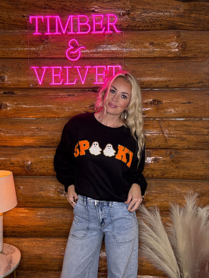 Person wearing a black sweater with 'SPooky' print sweatshirt in front of a wooden wall with a neon sign.