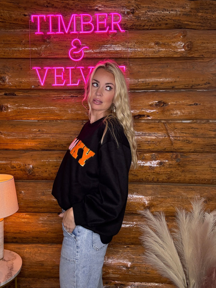 Person wearing a spooky sweatshirt standing in front of a wooden wall with a neon sign reading 'Timber & Velvet'.