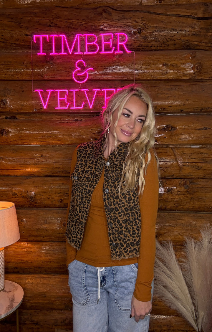 Person standing in front of a wooden wall with a neon sign reading 'Timber & Velvet' wearing a  leopard vest.