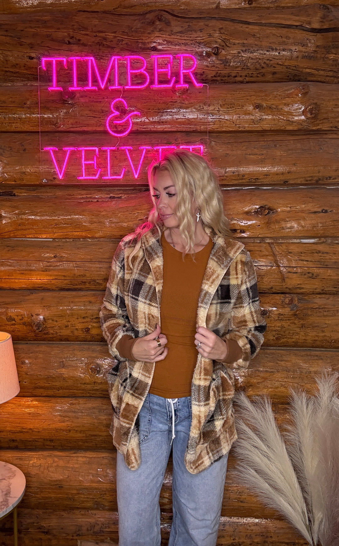 Woman in a plaid jacket standing in front of a wooden wall with a neon sign reading 'Timber & Velvet'.