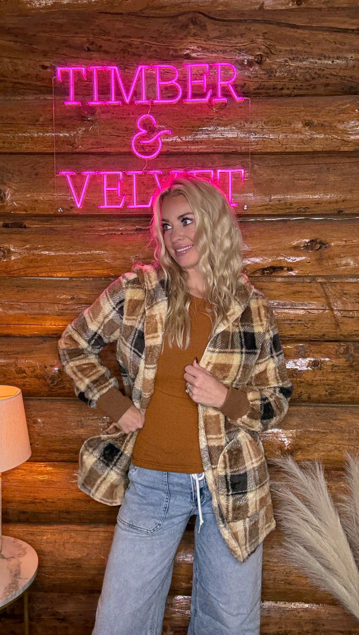 Person wearing a plaid jacket in front of a wooden wall with 'Timber & Velvet' neon sign.