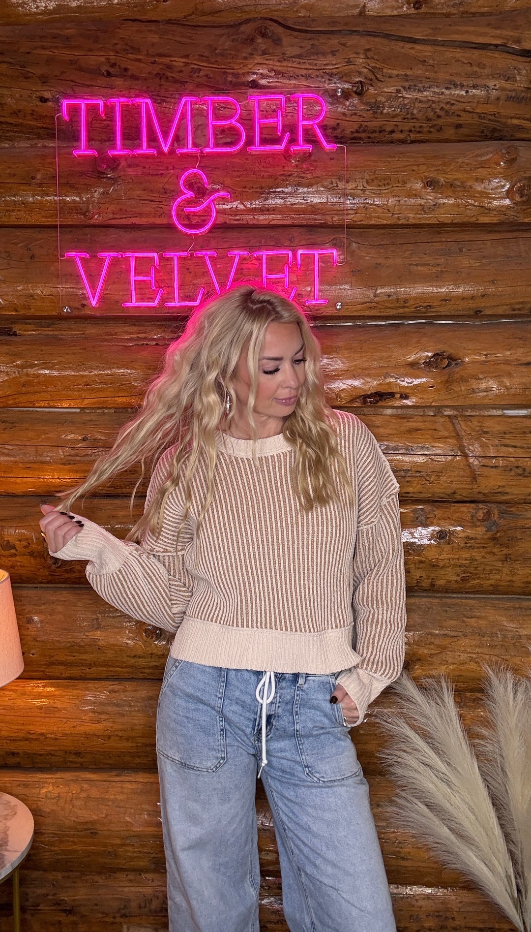 Person standing in front of a wooden wall with a neon sign reading 'Timber & Velvet'. Wearing a two toned sweater in cream and brown