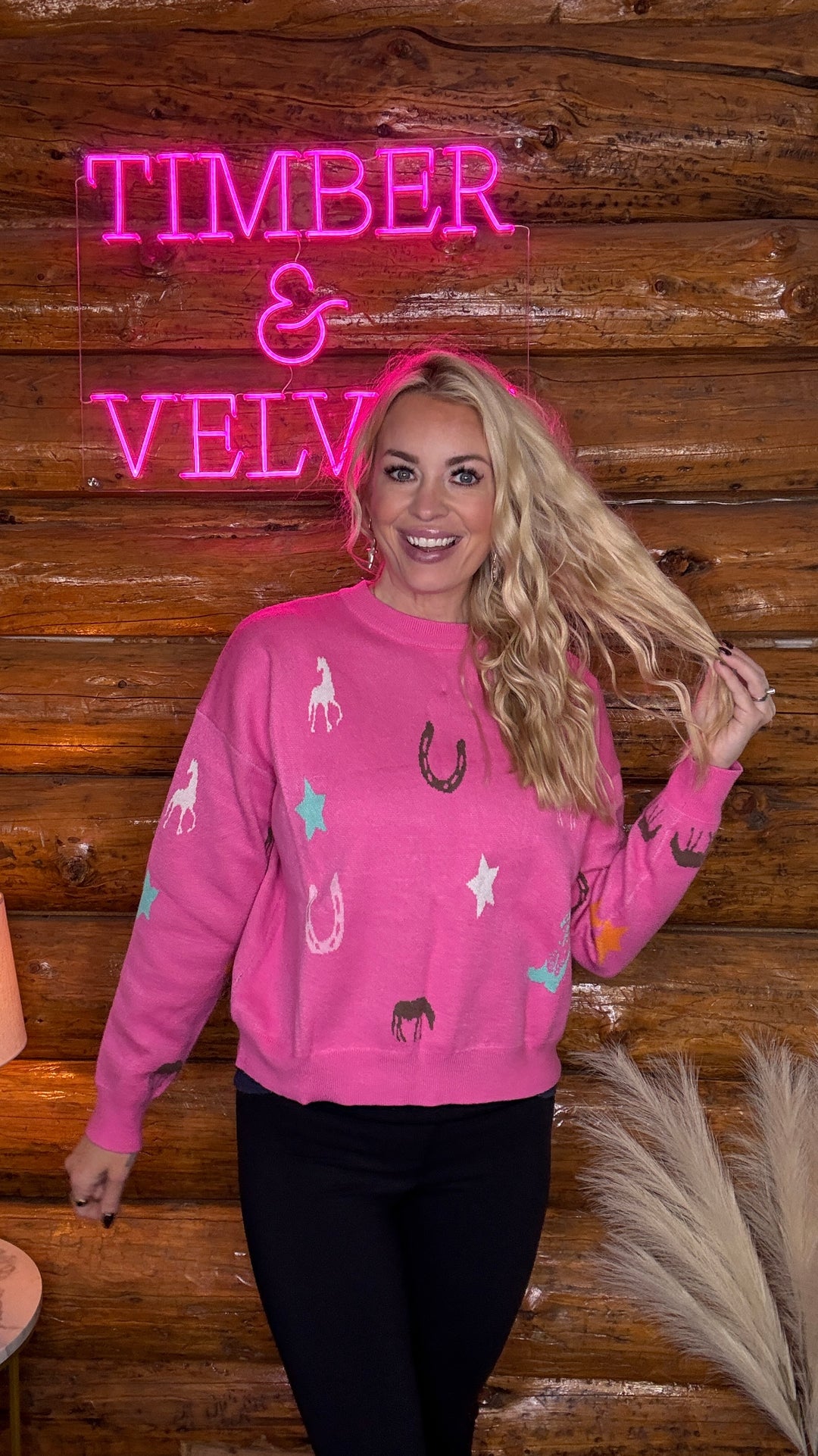 Person wearing a pink sweatshirt with horse designs in front of a wooden wall with 'Timber & Velv' neon sign.
