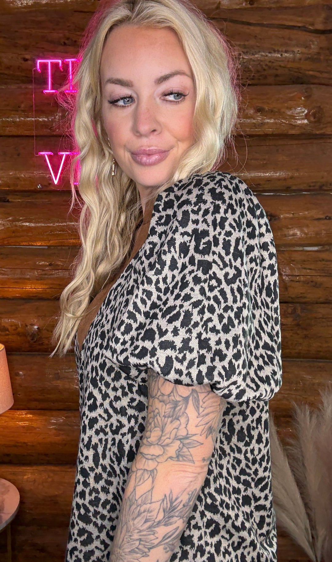 Person wearing a leopard print outfit with tattoos visible, standing against a wooden wall with neon signs.