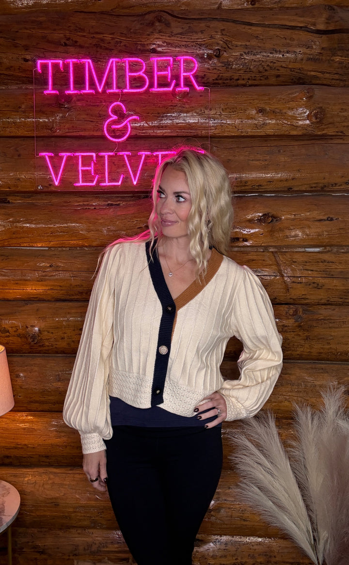 Woman standing in front of a wooden wall with 'Timber & Velvet' neon sign wearing a color block cardigan 