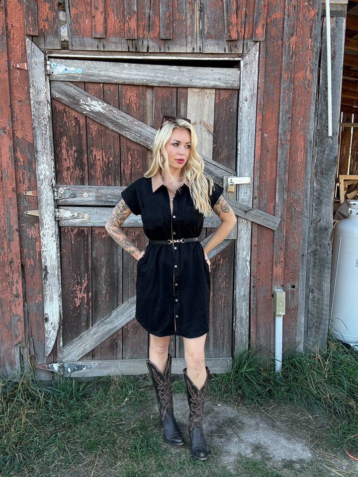 Woman in a black button down shirt dress and boots standing in front of a rustic wooden door.