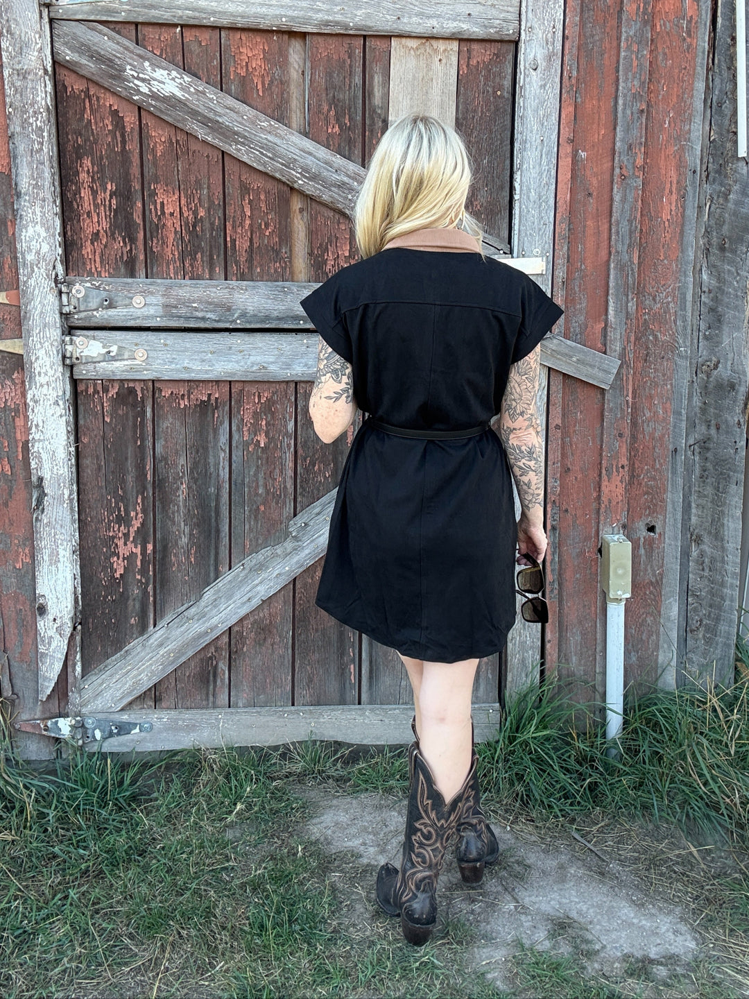Person wearing a black button down shirt dress and boots standing in front of a rustic wooden door.t 