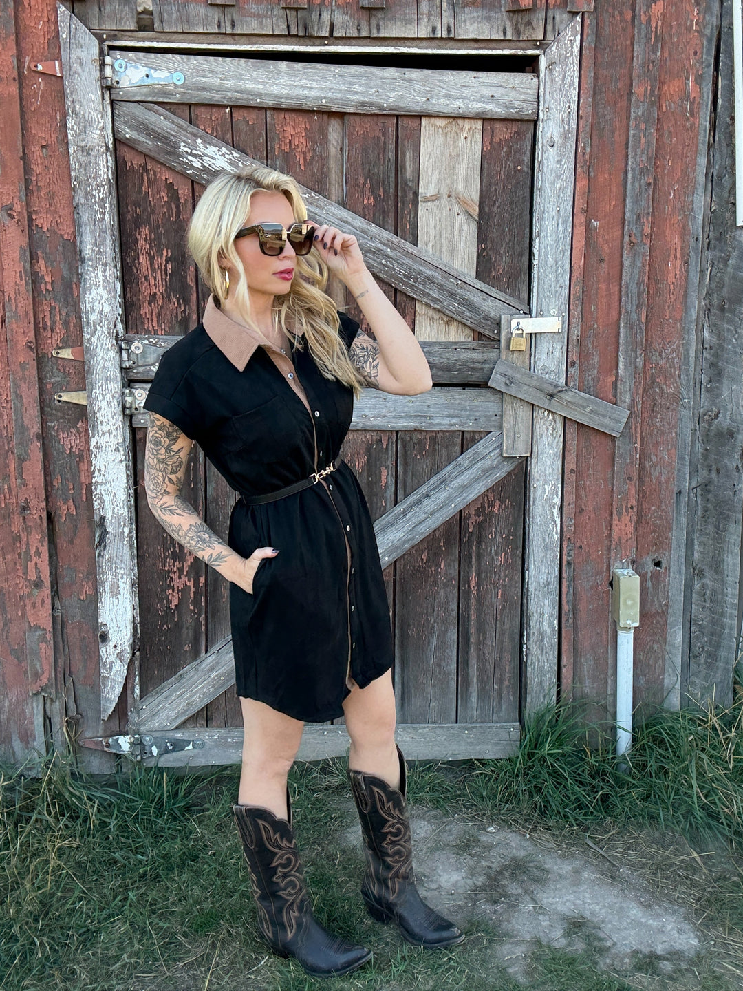 Woman in a black button up dress and cowboy boots standing in front of a rustic wooden door.