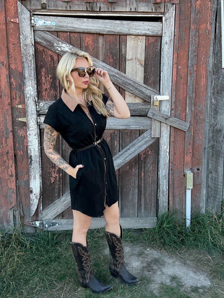 Woman in a black button up dress and cowboy boots standing in front of a rustic wooden door.