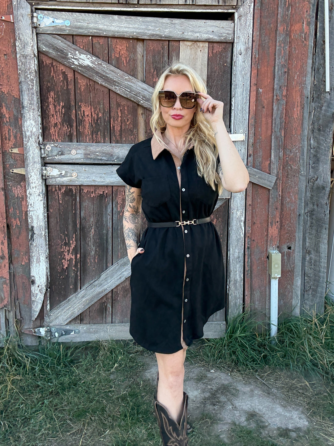 Woman in a black button down shirt dress standing in front of a rustic wooden door.
