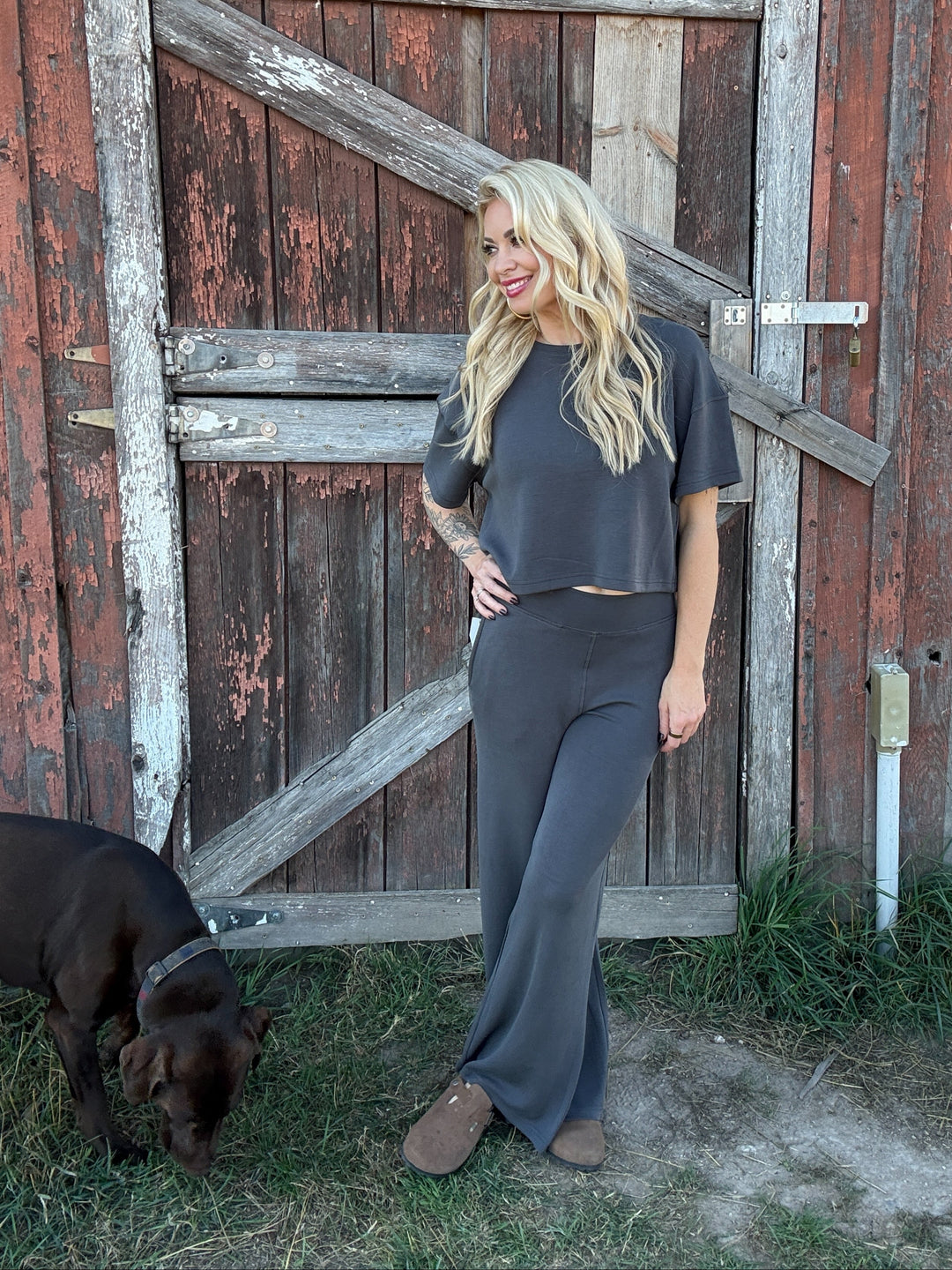 Woman in gray lounge outfit standing next to a chocolate lab dog in front of a rustic wooden door.