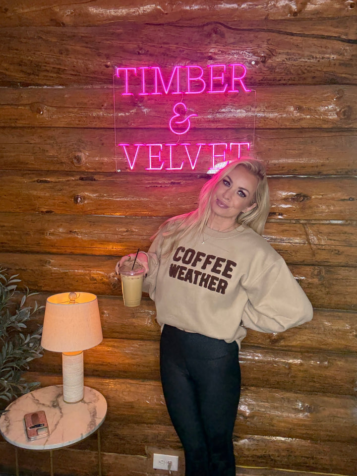 Woman holding a drink wearing a coffee weather sweatshirt in front of a neon sign with 'Timber & Velvet' on a wooden wall.