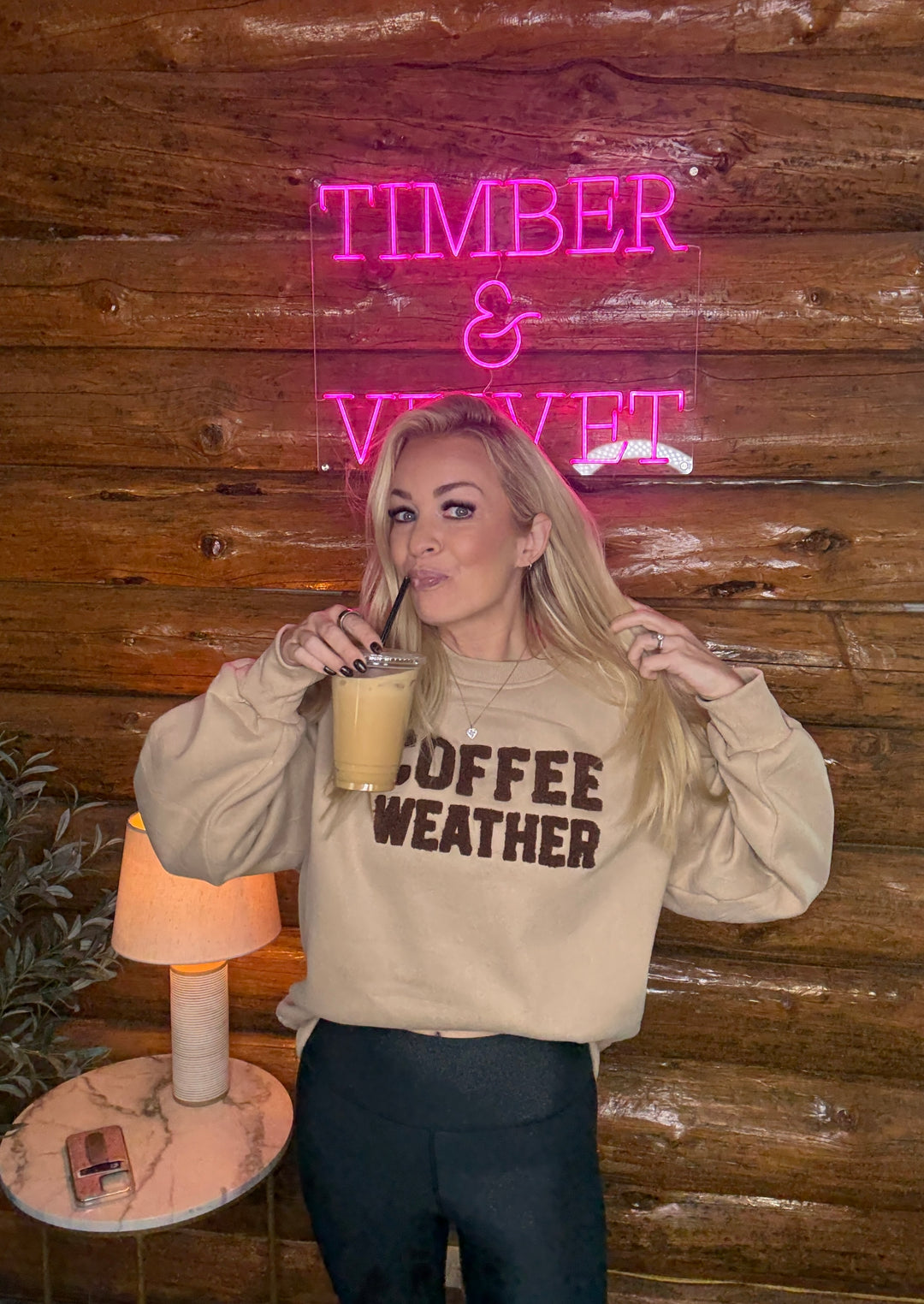 Person wearing a 'Coffee Weather' sweatshirt in front of a neon sign with a wooden wall background.
