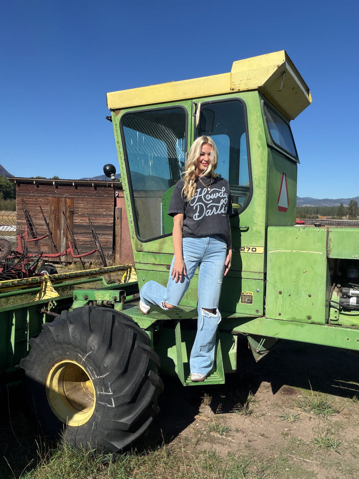 Person wearing a 'Howdy Darlin'' Tee standing on a large green tractor with a clear blue sky in the background