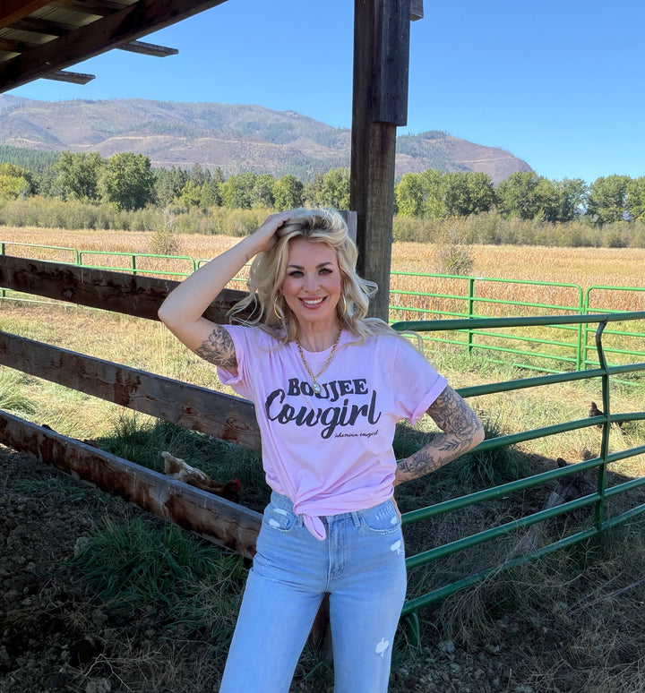 Woman wearing a 'BOJEE Cowgirl' t-shirt standing in a rural setting with mountains in the background.