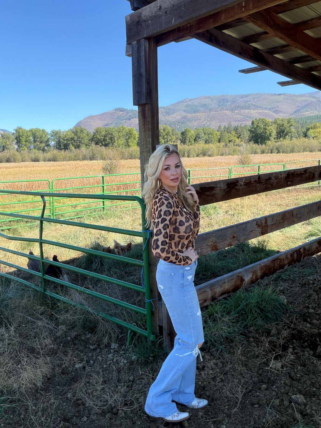 Person wearing a leopard print mesh top and white pants standing near a wooden structure with a scenic background.