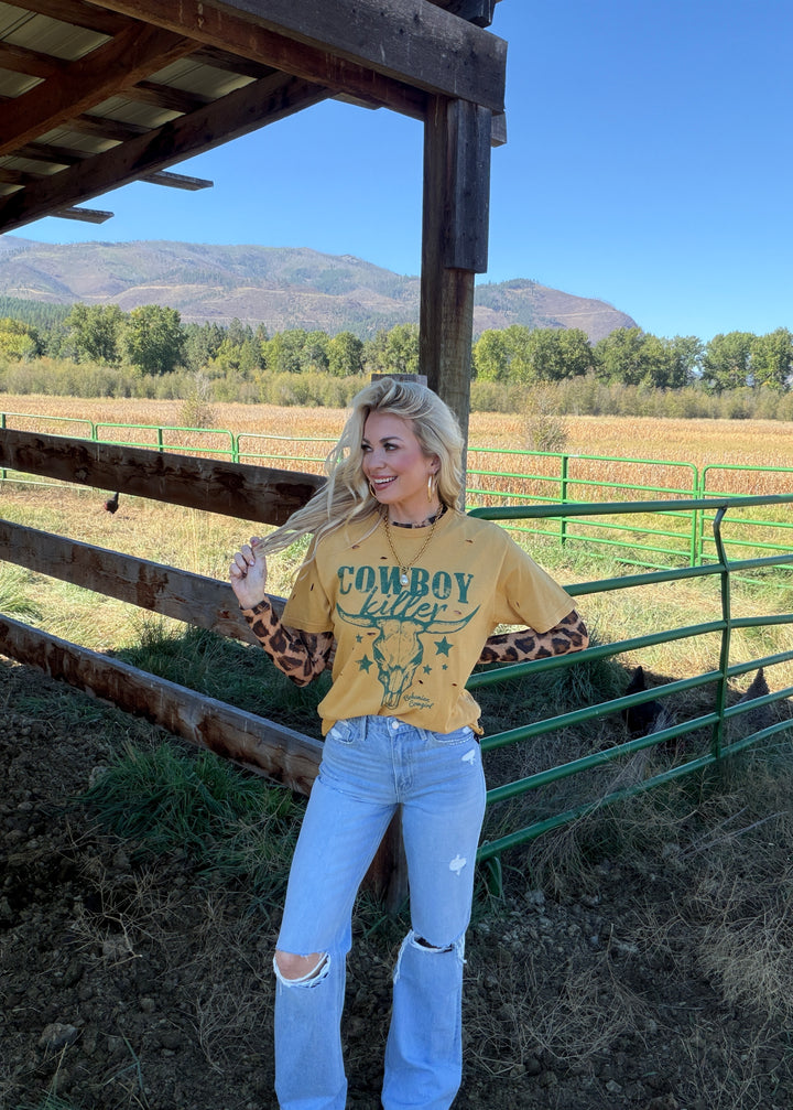 Woman wearing a mustard yellow 'Cowboy Killer' shirt with leopard print sleeves in a rural setting.
