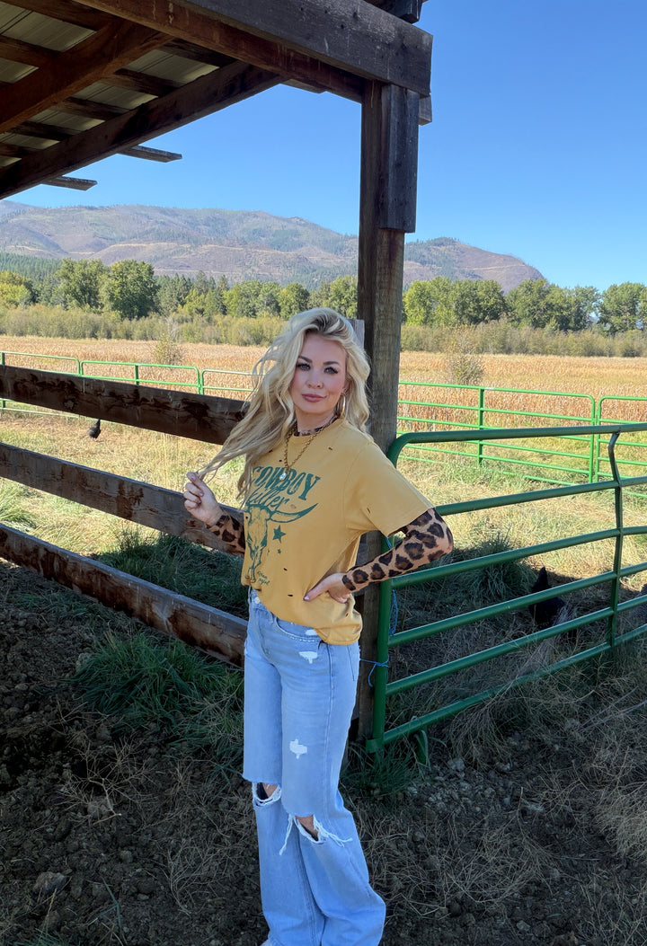 Woman in a yellow 'Cowboy Killer' shirt and light blue jeans standing in a field with mountains in the background