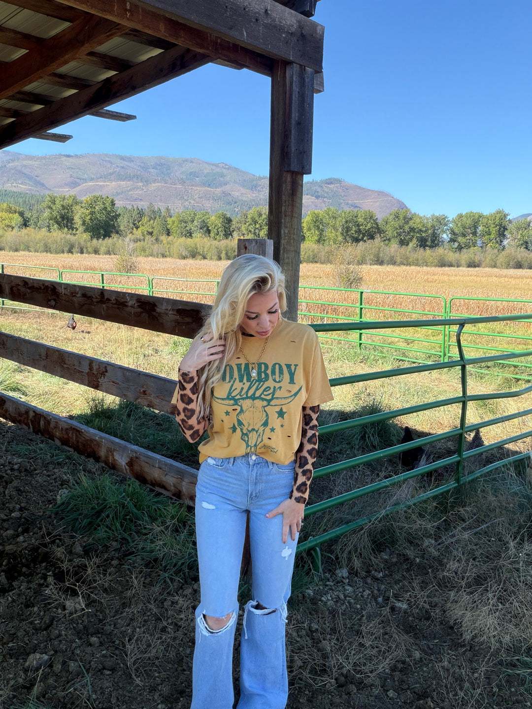 Woman wearing a yellow long-sleeve shirt with 'Cowboy Killer' text and blue jeans standing in a barn.