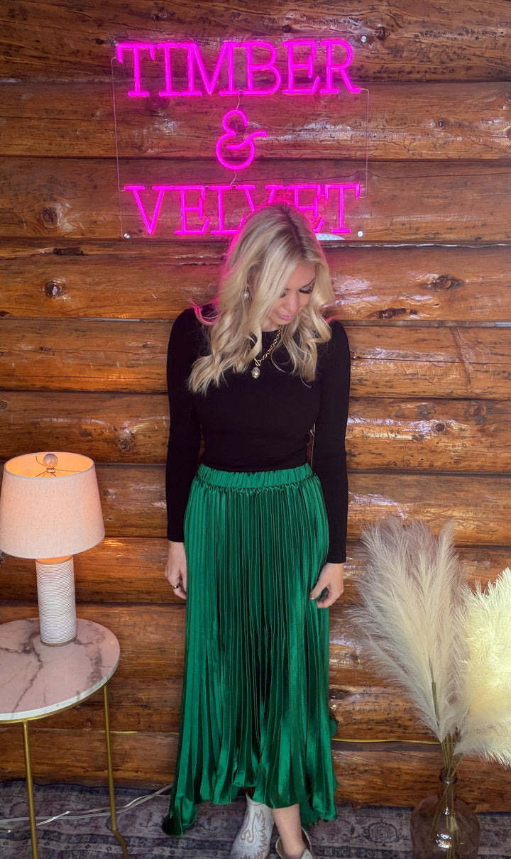 Woman in a black top and green pleated skirt standing in front of a wooden wall with 'Timber & Velvet' neon sign.
