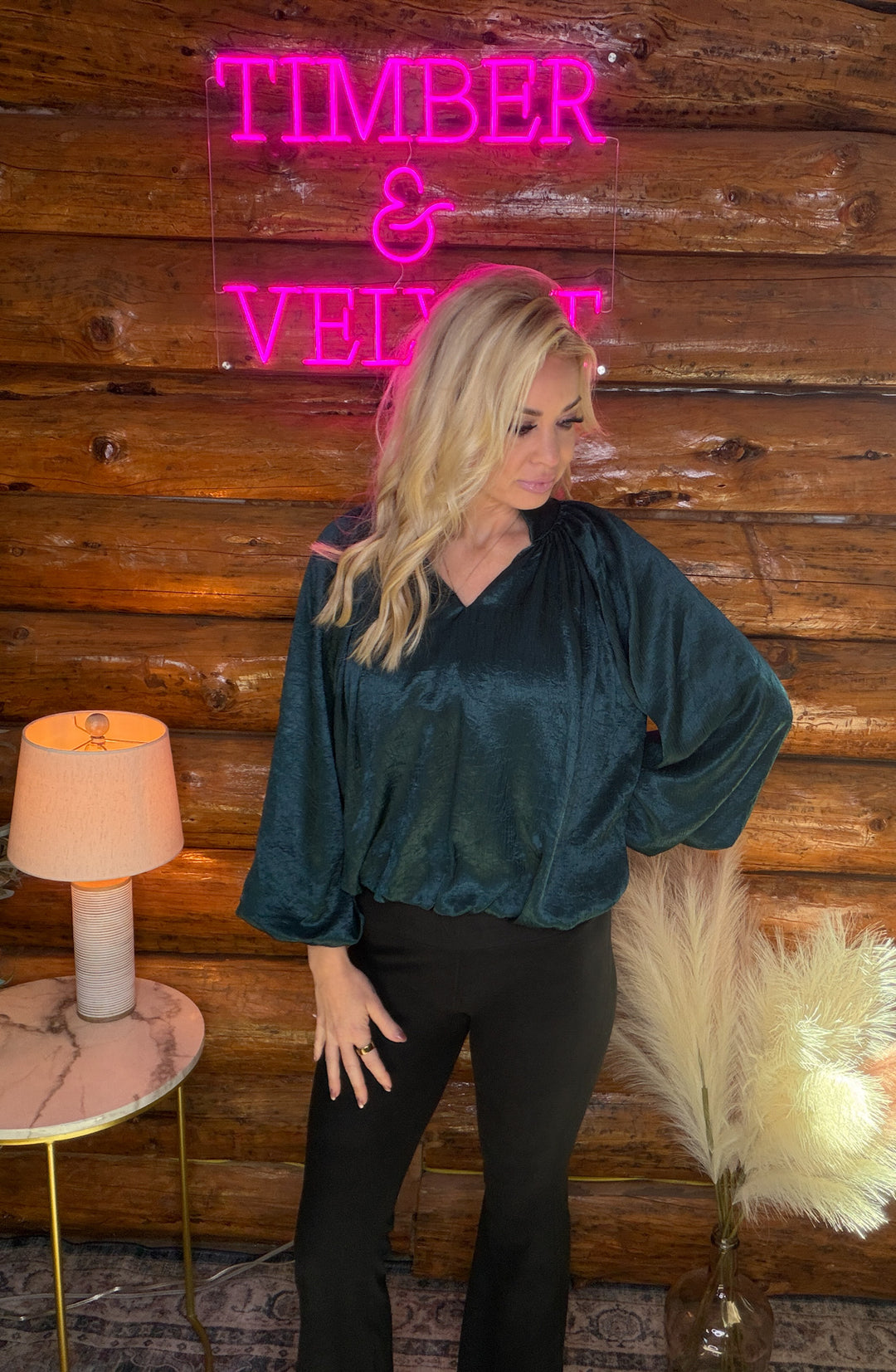 Woman wearing a green holiday blouse posing in front of a wooden wall with a neon sign and decorative elements.