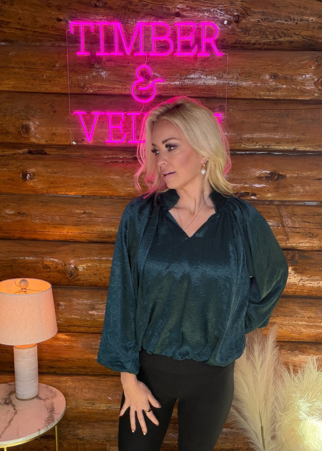 Woman wearing a green holiday blouse standing in front of a wooden wall with a neon sign reading 'Timber & Vine'.