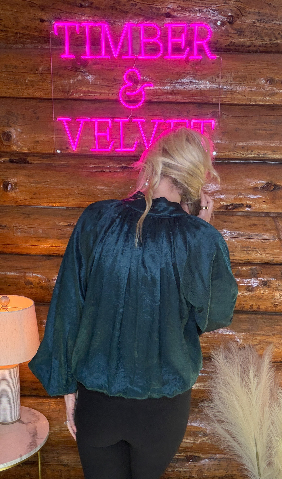 Person wearing a teal blouse in front of a wooden wall with a neon sign reading 'TIMBER & VELVET'.