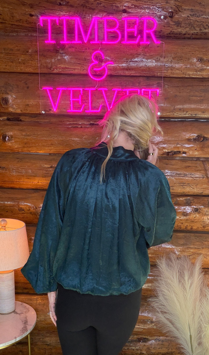 Person wearing a teal blouse in front of a wooden wall with a neon sign reading 'TIMBER & VELVET'.