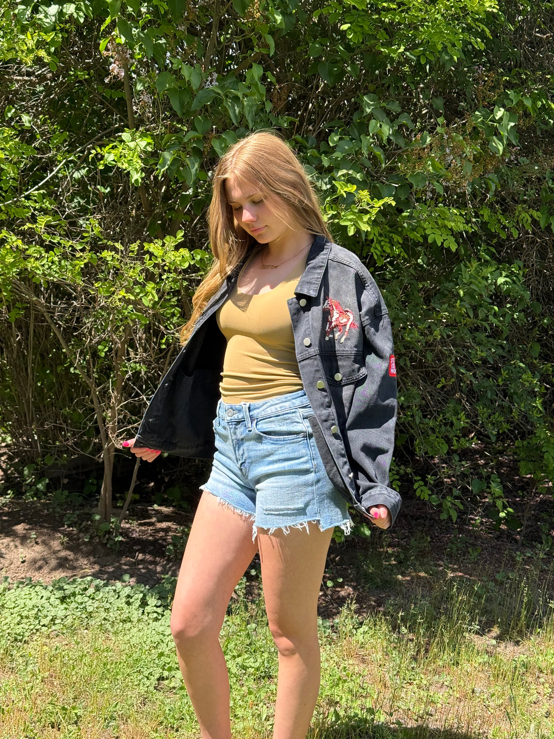 Woman wearing a denim jacket, yellow top, and denim shorts standing outdoors with greenery in the background