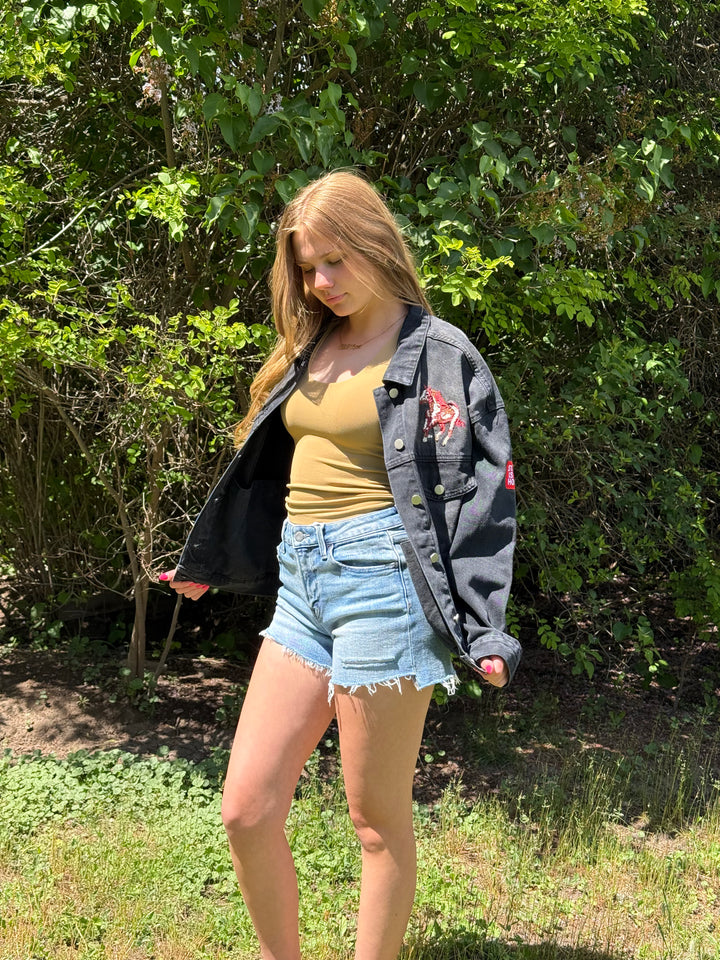 Woman wearing a denim jacket, yellow top, and denim shorts standing outdoors with greenery in the background