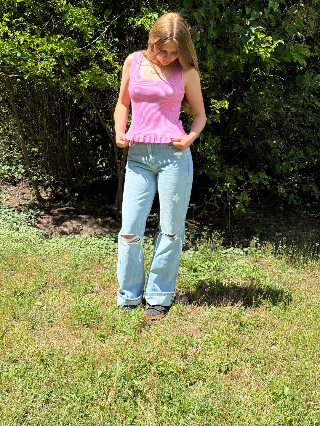 Woman in pink top and blue jeans standing in a grassy area with greenery.