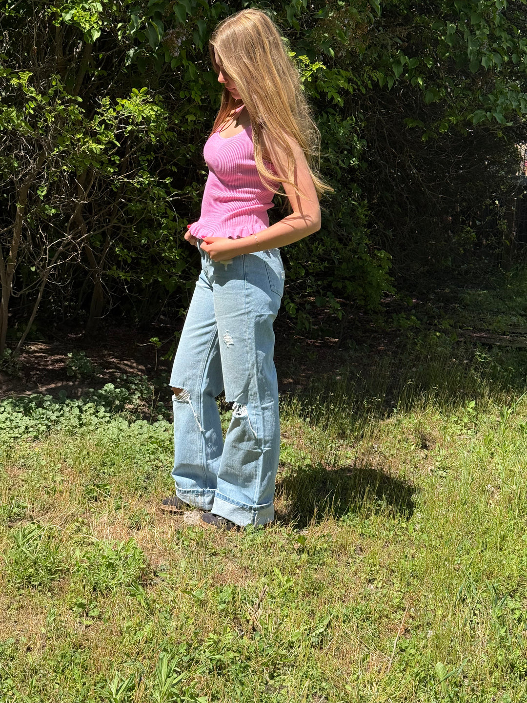 Woman in a pink top and blue jeans standing in a grassy area with trees in the background