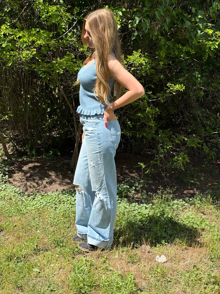 Woman in a light blue sleeveless top and jeans standing outdoors with greenery in the background