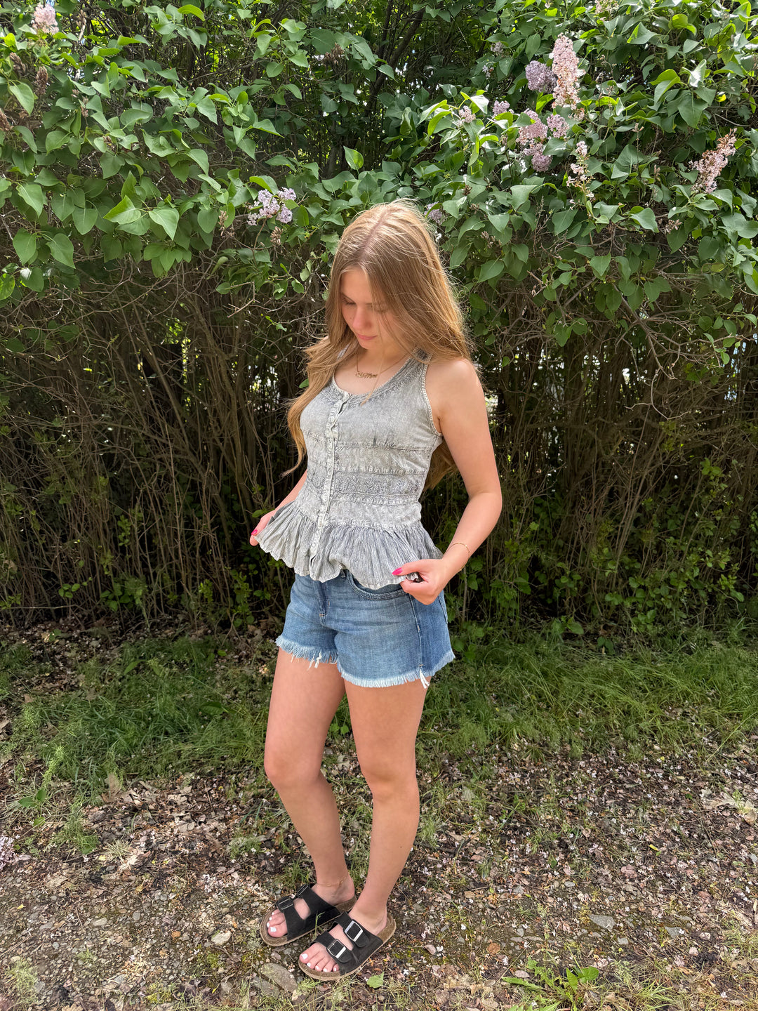 Woman wearing a light blue sleeveless top and denim shorts standing outdoors with greenery in the background.