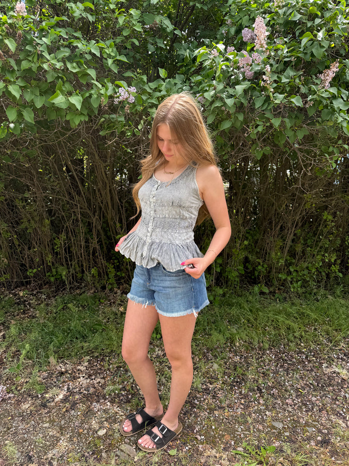 Woman wearing a light blue sleeveless top and denim shorts standing outdoors with greenery in the background.