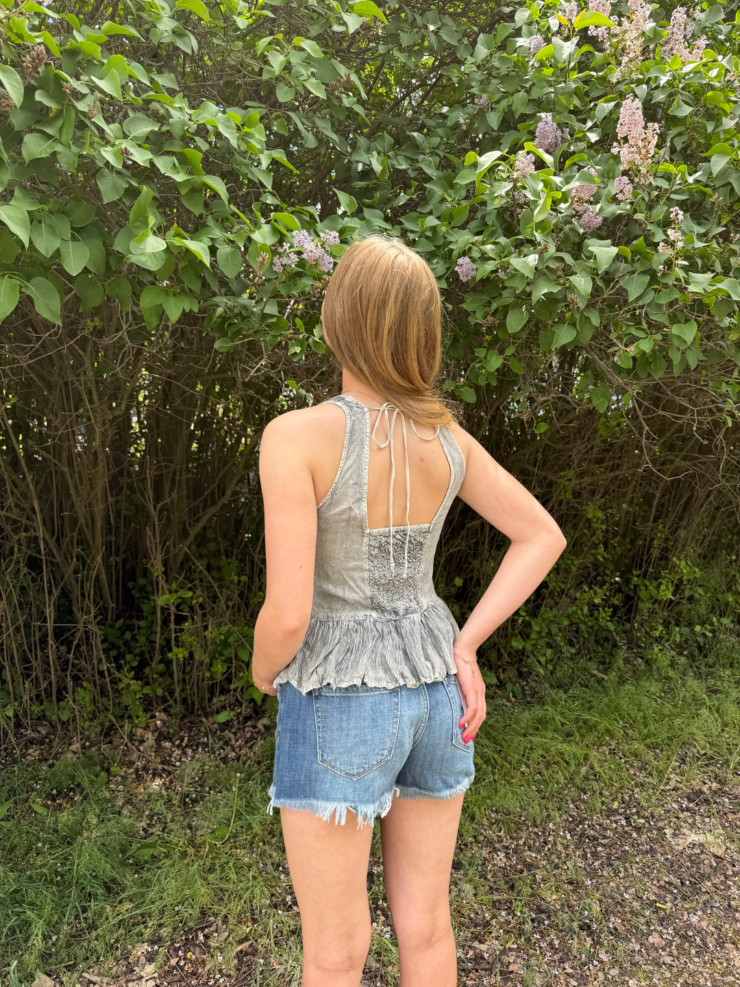 Person wearing a grey sleeveless top and denim shorts standing in front of a bush with purple flowers.