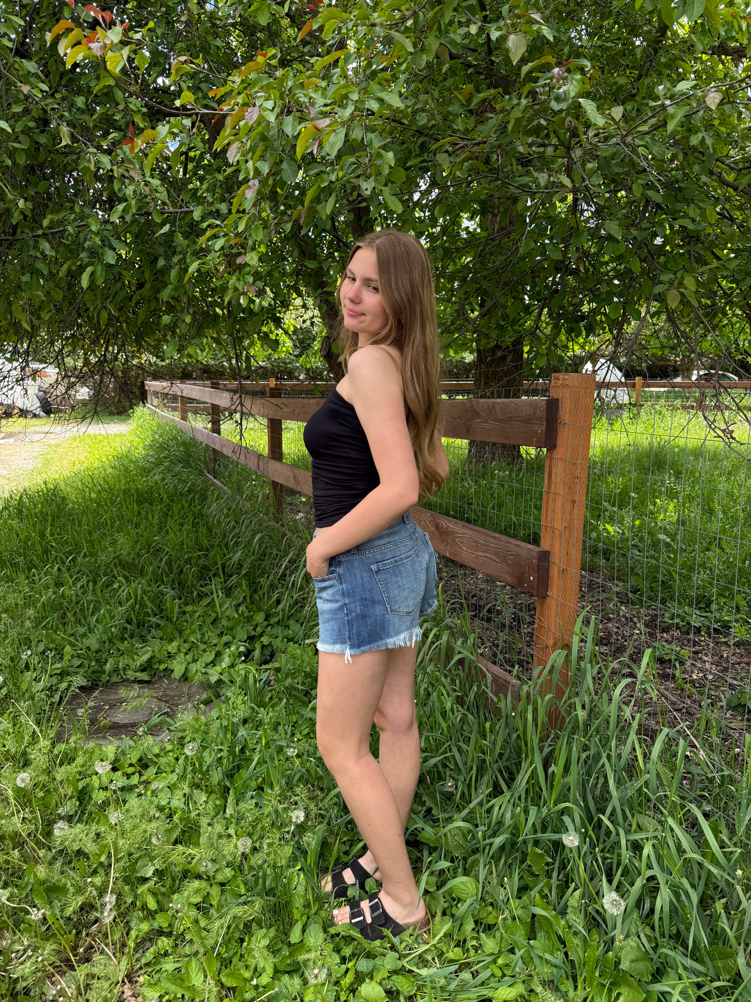 Woman wearing a black tube top and shorts standing in a grassy area with a wooden fence and trees in the background