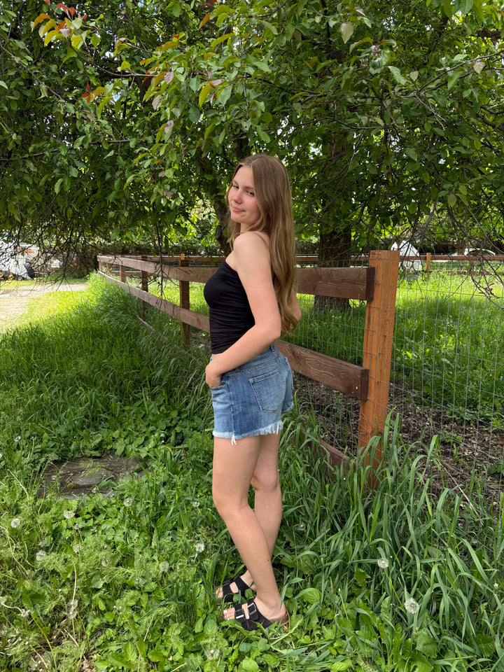 Woman wearing a black tube top and shorts standing in a grassy area with a wooden fence and trees in the background