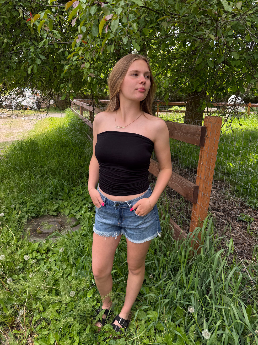 Woman in a black tube top and denim shorts standing in a grassy area with trees and a wooden fence in the background.