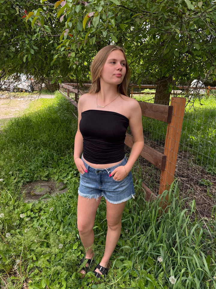 Woman in a black tube top and denim shorts standing in a grassy area with trees and a wooden fence in the background.