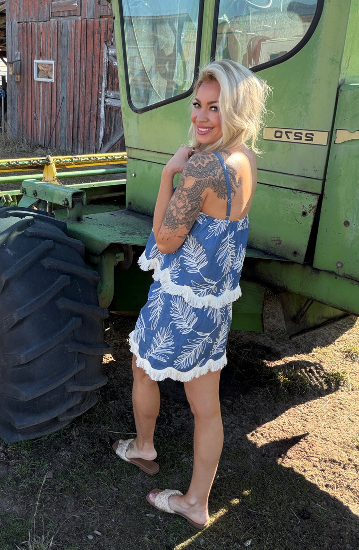 Woman in a blue top and skort with embroidered leaves standing next to a green tractor on a sunny day.