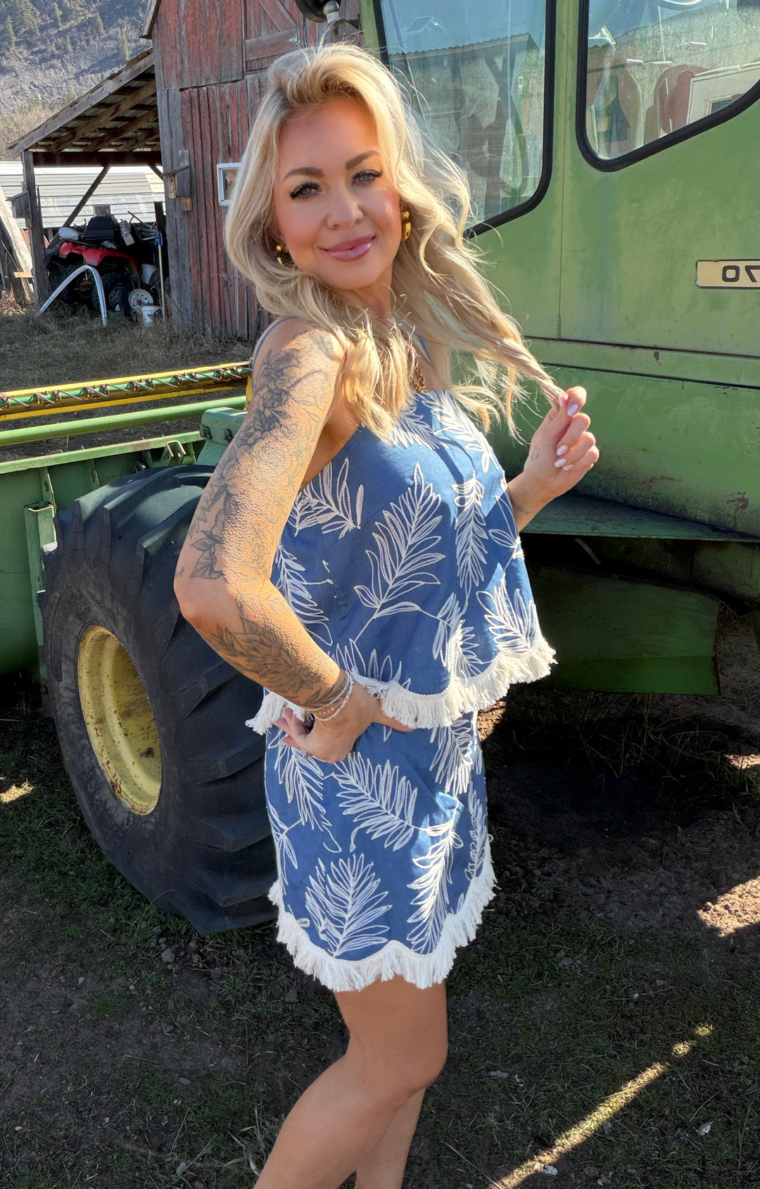 Woman in a blue tank & skort standing in front of a green tractor on a farm.