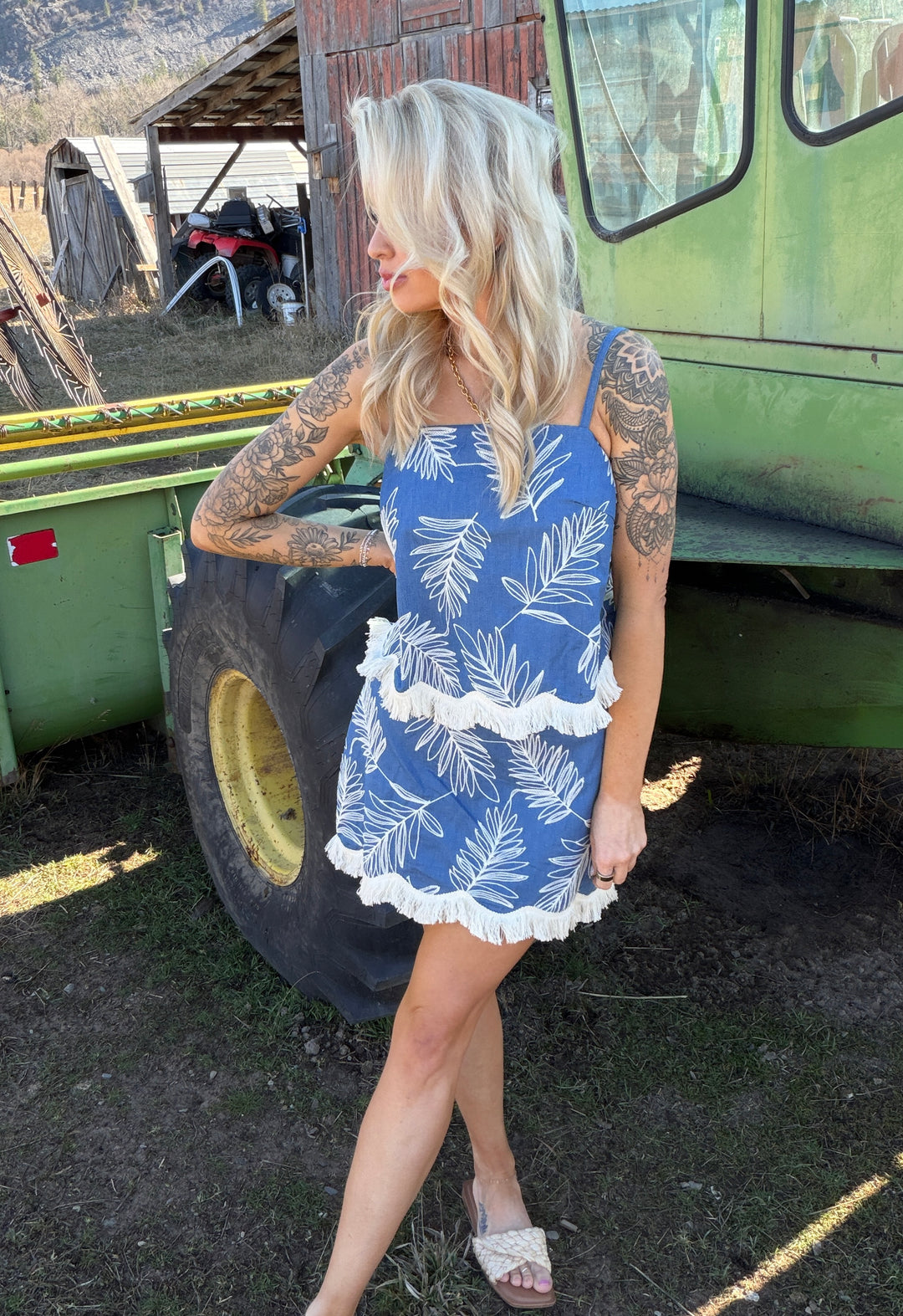 Woman in a blue top & skort with white leaf pattern standing next to a green tractor on a farm.