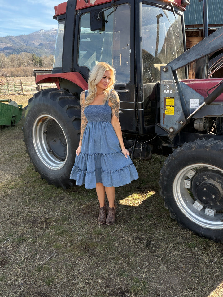 Woman in a blue denim dress standing next to a large tractor on a dirt field.