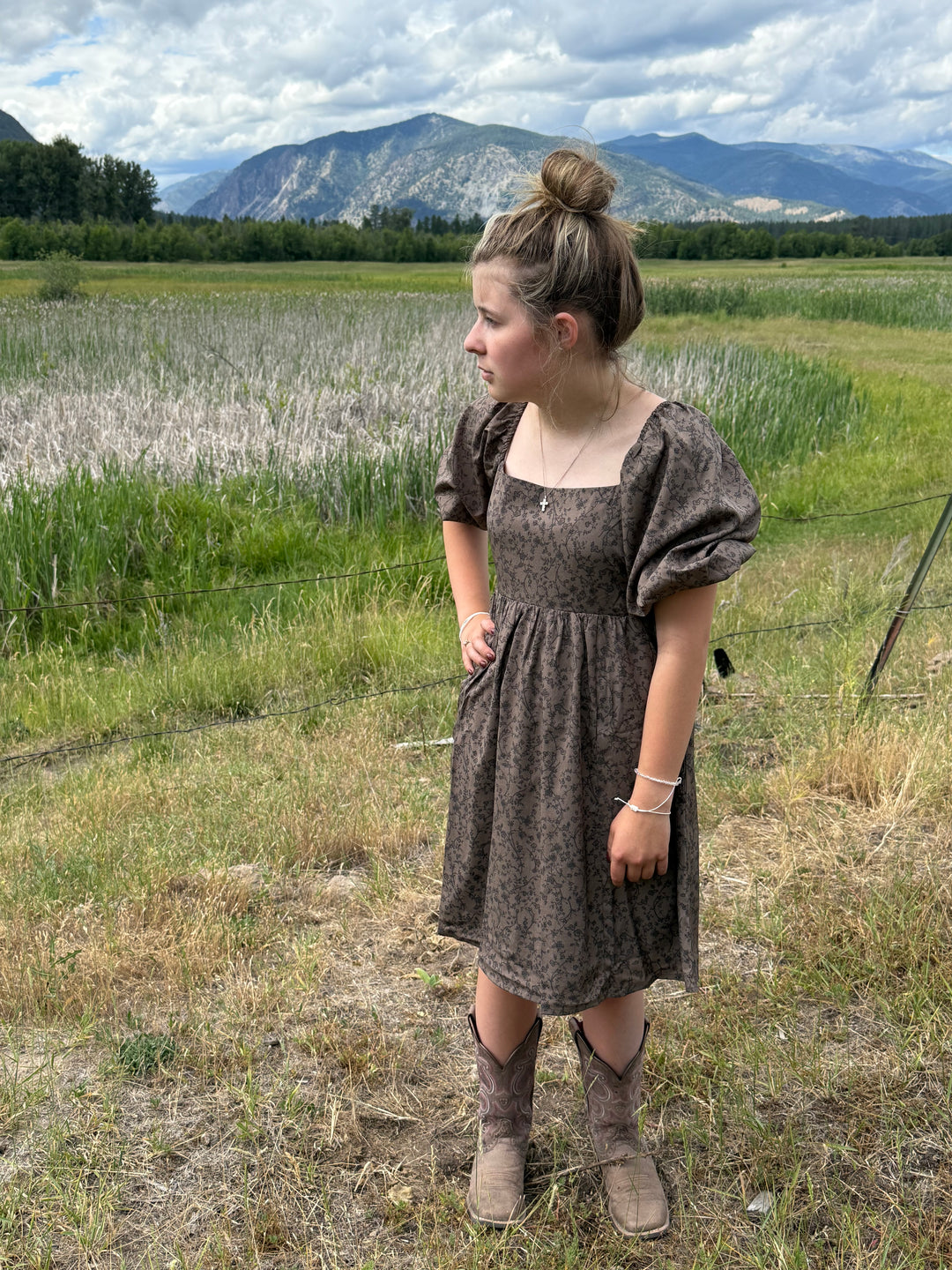 Person in a brown floral dress standing in a grassy field with mountains in the background