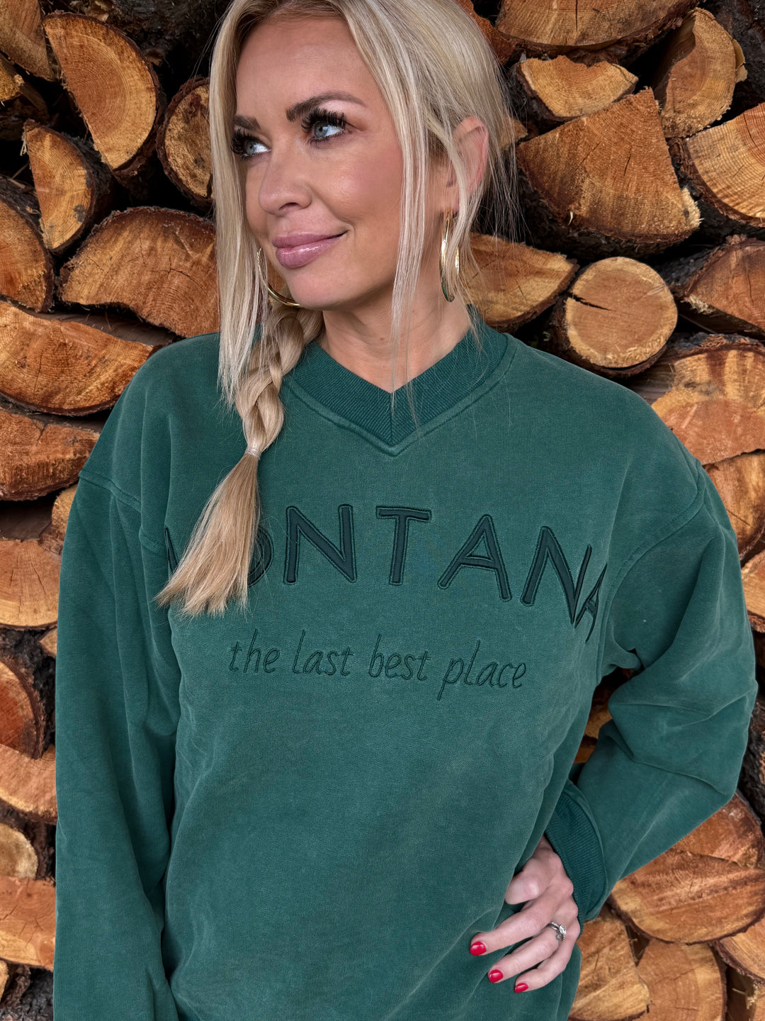 Person wearing a Montana green sweatshirt with the last best place text, standing among stacked wooden logs.