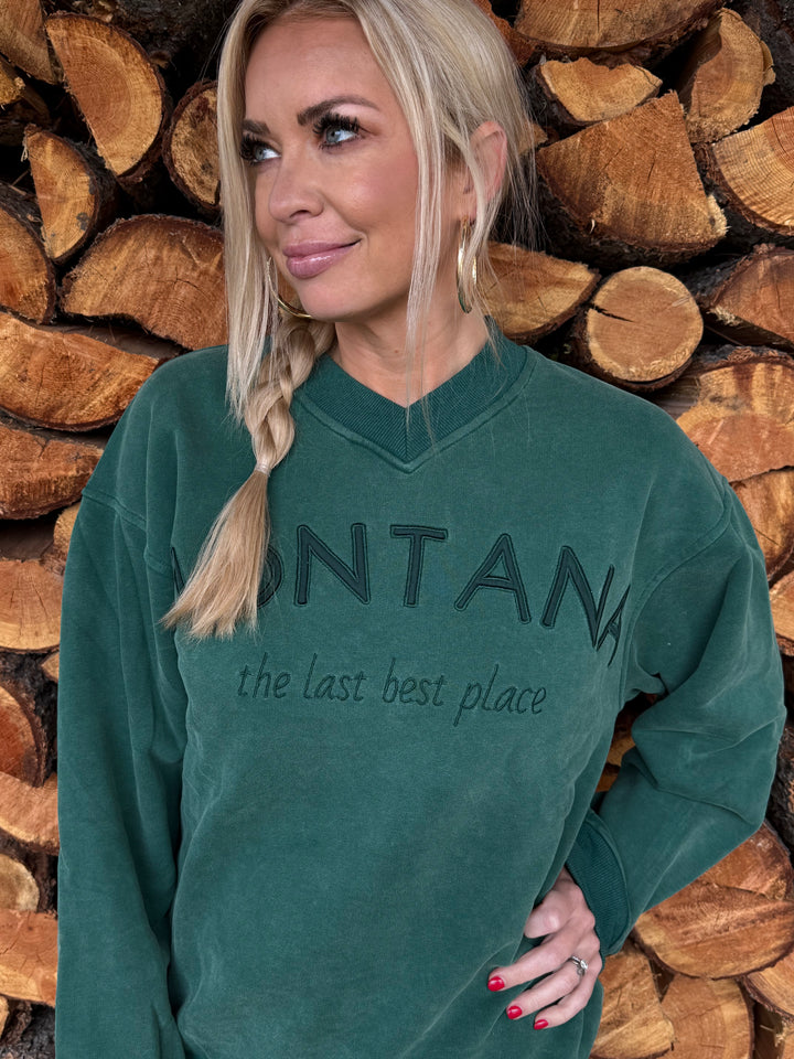 Person wearing a Montana green sweatshirt with the last best place text, standing among stacked wooden logs.