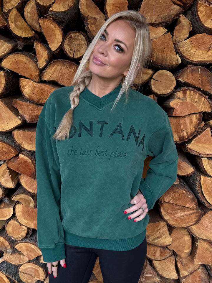 Woman wearing a green sweatshirt with 'Montana' text, standing among stacked wooden logs.