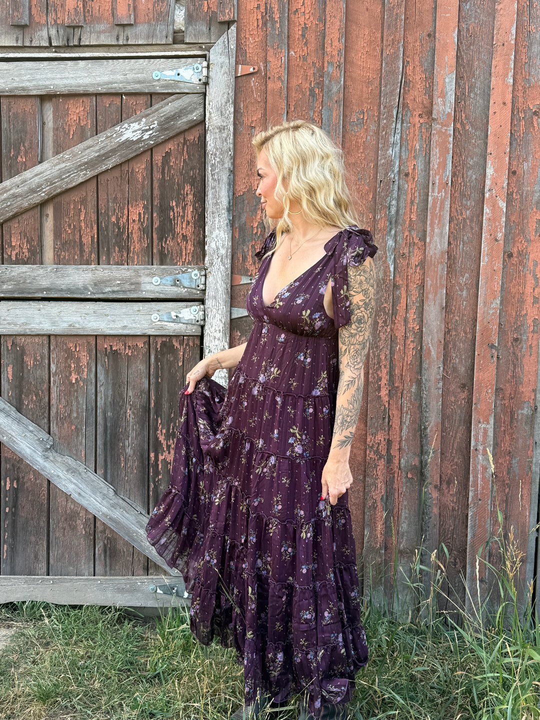 Woman in a long dark purple flower dress standing next to a wooden barn.