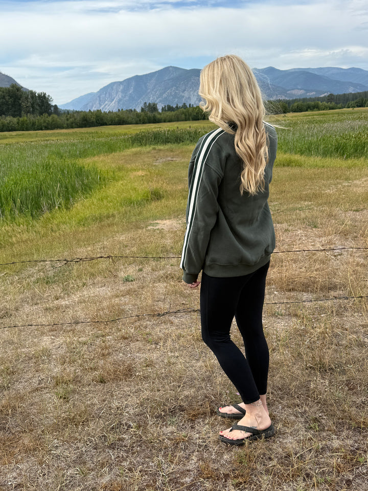 Person standing on a dirt path wearing a green Montana crewneck with a scenic view of mountains and fields.